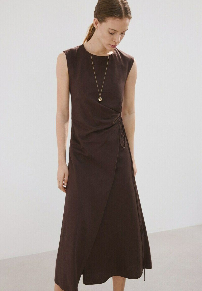 Femeie purtând o rochie înfășurată fără mâneci, de culoare maro închis, cu legături laterale, privind în jos, pe un fundal simplu gri deschis.