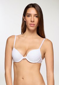 Reggiseno bianco in pizzo con coppe sagomate, spalline regolabili e un centro decorativo, caratterizzato da motivi floreali e una texture liscia.
