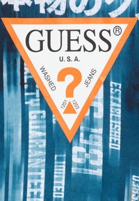 Tela de denim azul y blanco desgastada con rayas verticales. Presenta un prominente logo triangular naranja con el texto: "GUESS U.S.A. WASHED JEANS."