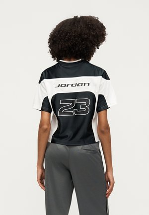 Osoba z kręconymi włosami ubrana w czarno-biały sportowy T-shirt Jordan z numerem 23 na plecach oraz szare spodnie.