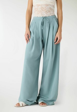 Donna con pantaloni a gamba larga, vita alta, di colore azzurro chiaro con coulisse e sandali bianchi, abbinati a un top corto in pizzo bianco.