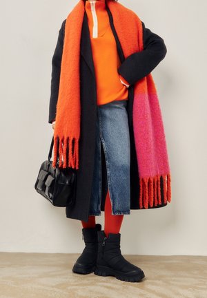 Person iført sort lang frakke, lys orange og pink tørklæde, orange top, blåt denimnederdel, røde strømpebukser, sorte støvler, holder en sort håndtaske.