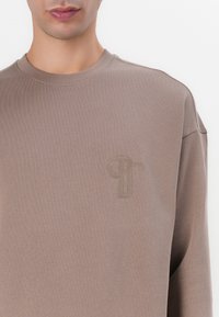 Sudadera marrón de tela suave, con un cuello redondo y un sutil logotipo embossado en la parte izquierda del pecho.