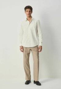 Cămașă crem cu butoane, cu un model subtil de puncte, un singur buzunar pe piept și mâneci lungi, asortată cu pantaloni bej și pantofi negri.