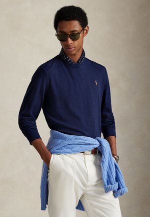 Homme portant une chemise à manches longues bleu foncé, un pantalon blanc, des lunettes de soleil et un pull bleu clair noué autour de la taille, debout les mains dans les poches.