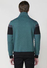 Sudadera con cremallera en color teal y cuello alto, con paneles de acento en negro y gris, y puños y cintura con canalé. Tejido texturizado.