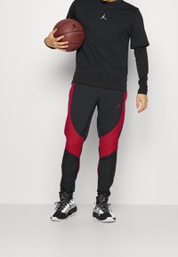 Camiseta negra de manga larga con logo, combinada con joggers negros y rojos con paneles curvados. Llevando zapatillas deportivas black y white, sosteniendo un balón de baloncesto.