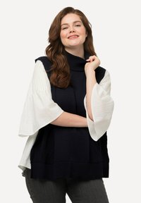 Top senza maniche lavorato a maglia di colore blu navy con colletto alto, abbinato a una blusa bianca con maniche svasate. Presenta motivi testurizzati e una vestibilità ampia.