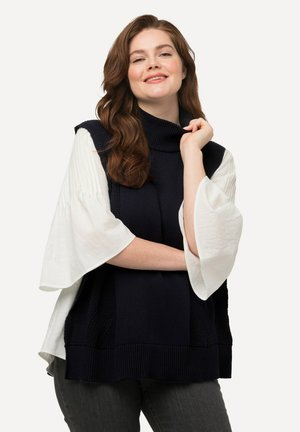 Top senza maniche lavorato a maglia di colore blu navy con colletto alto, abbinato a una blusa bianca con maniche svasate. Presenta motivi testurizzati e una vestibilità ampia.