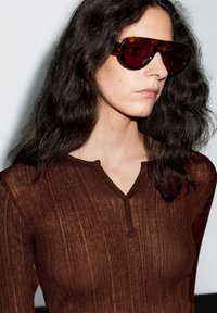 Hnědý žebrovaný top s dlouhým rukávem a výstřihem do V, doplněný o velké sluneční brýle s želvovinovým vzorem a tmavými čočkami.