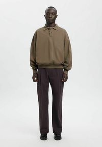 Brūns pārmēru polo krekls ar noslīpiem pleciem, kombinēts ar tumši pelēkām plaša izgriezuma biksēm. Audums izskatās mīksts ar gludu tekstūru.