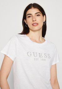 Balta trumpa rankovė t-shirt marškinėliai iš lengvos medžiagos. Su "GUESS" logotipu ir "EST. 1981" studiniu akcentu. Lygus paviršius.
