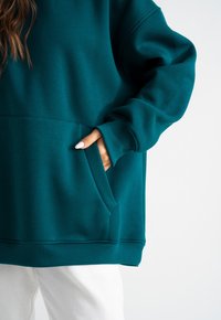 Felpa oversized color turchese con tasca frontale, polsini e orlo a coste. Il tessuto sembra morbido e strutturato, adatto per un look casual.