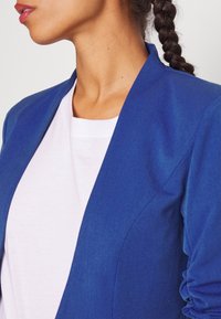Femme aux cheveux tressés portant une chemise blanche sous un blazer bleu, visible du cou jusqu'au milieu du torse sur un fond uni.