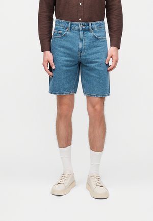 Férfi kék farmer rövidnadrágot, fehér zoknit, bézs tornacipőt és barna hosszú ujjú inget visel, fehér háttér előtt.