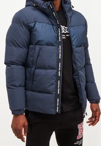 Tmavě modrá prošívaná bunda s kapucí, opatřená zipem a páskem s logem podél zipu. Hladká textura a elastické manžety.