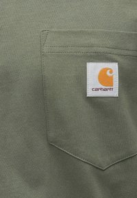 Olivově zelené tričko s náprsní kapsou; má bílou etiketu s oranžovým logem a textem. Měkká, strukturovaná bavlněná tkanina s pevným šitím.