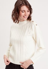 Un pull en tricot couleur crème avec un col montant, des torsades texturées, des motifs en diamant et des accents de boutons dorés sur l'épaule.