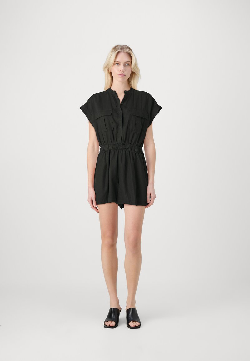 Combinaison noire à manches courtes avec un devant boutonné, deux poches poitrine, taille élastique et coupe ample. Associée à des chaussures glissantes noires.