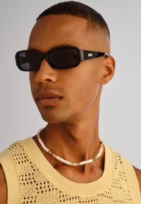 Νέος άνδρας με κοντά μαλλιά που φοράει μαύρα ορθογώνια γυαλιά ηλίου, μπεζ πλεκτό μπλουζάκι χωρίς μανίκια και λευκό κολιέ με χάντρες.