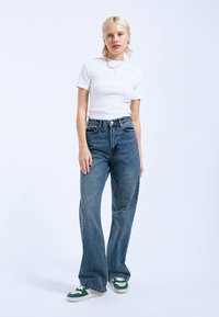 Hoge taille blauwe spijkerbroeken met een losse pasvorm, gecombineerd met een aansluitend wit T-shirt en sneakers met groene accenten. Eenvoudige juweeldetails.