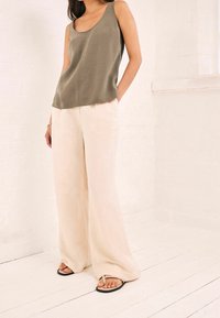 Donna che indossa un top senza maniche verde oliva, pantaloni larghi crema e sandali flip-flop neri, in piedi in una stanza chiara.