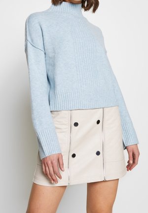 Mujer con suéter de cuello alto de canalé azul claro y minifalda crema con botones negros y cremalleras verticales.