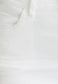 Gros plan sur un tissu en denim blanc montrant le bord d'une poche avant, une boucle de ceinture et des coutures détaillées.