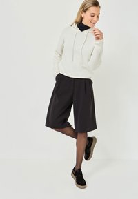 Vit fleece hoodie med dragsko, kombinerad med svarta veckade shorts. Modellen bär svarta sneakers med texturerade sulor.