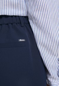 Blå och vit randig skjorta instoppad i marinblå byxor med elastisk midja och en liten silverlogotyp nära fickan.