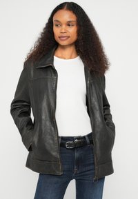 Jachetă de piele neagră cu fermoar frontal, guler, două buzunare laterale și cusături contrastante. Purtată peste o cămașă albă și blugi albaștri.