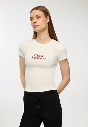 T-shirt bianca aderente con maniche corte, caratterizzata da testo rosso "Ciao Amore" sul petto. Il testo presenta un design leggermente curvo.