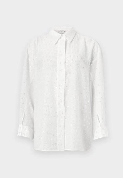 Cămașă albă cu mâneci lungi, cu nasturi, cu un model subtil punctat din paiete și guler clasic pe fundal simplu.
