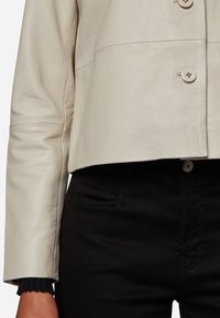 Ljusbeige läderkorta jacka med knappar, med en slät textur och sömdetalj. Matchad med svarta byxor, som visar fickor och sömmar.