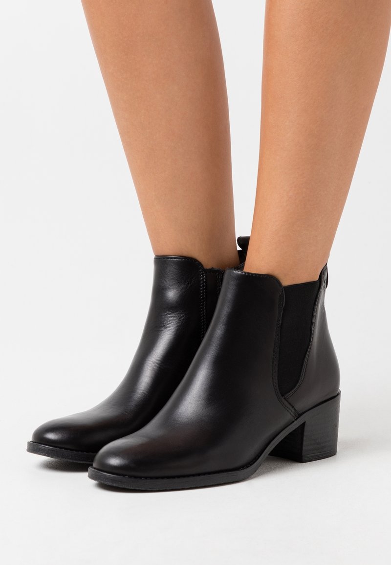 Bottes pour cheville en cuir noir avec talons bas empilés portées sur des jambes nues, avec des panneaux latéraux élastiques et des bouts arrondis.