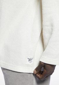 Dłoń trzyma brzeg białego swetra Reebok z prążkowanej dzianiny z etykietą barwioną naturalnie, noszonego z jasnoszarymi spodniami.