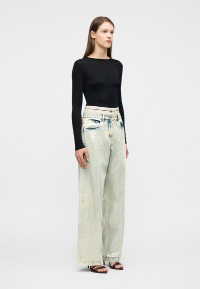 Γυναίκα στέκεται με το πρόσωπο προς τα δεξιά, φορώντας μαύρο μακρυμάνικο τοπ, ανοιχτόχρωμα τζιν με φαρδύ πόδι και μαύρα σανδάλια με τακούνια και λουριά.