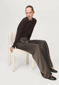 Model purtând un top maro închis, cu mâneci lungi, mulat, și pantaloni largi, cu talie înaltă, netezi, de nuanță maro metalic, așezată pe un scaun alb.