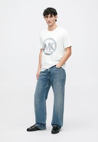 Tânăr îmbrăcat într-un tricou alb Michael Kors, blugi largi albaștri și adidași negri, cu o mână în buzunar.