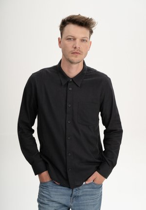 Junger Mann mit heller Haut und kurzen braunen Haaren trägt ein schwarzes Hemd mit Knöpfen und blaue Jeans, Hände in den Taschen, schaut ruhig nach vorne.