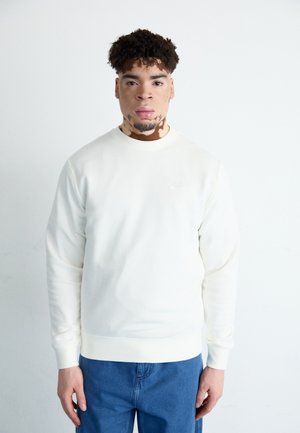 Sudadera crema con cuello redondo y mangas largas, con un pequeño logotipo bordado en el pecho. El modelo la combina con jeans azules.