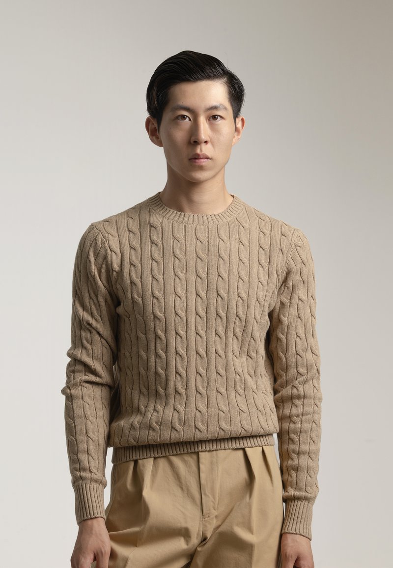 Suéter de punto beige con cuello redondo y puños de canalé, combinado con pantalones beige claros, que muestran patrones texturizados en toda su superficie.