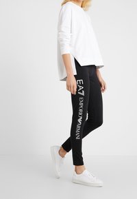 Svarta leggings med vit tryck av "EA7 Emporio Armani"-logotyp, ihop med en löst sittande vit sweatshirt och vita sneakers.