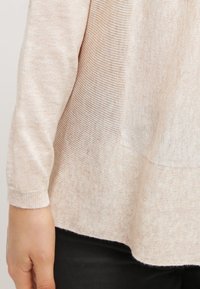 Jasnoróżowy sweter dziergany na drutach z długimi rękawami, luźnym krojem i teksturowanym wzorem, z zaokrąglonym dołem i subtelnymi różnicami w odcieniu.