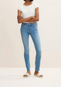 Top corto de manga corta blanco con borde ondulado, combinado con jeans ajustados de color azul claro y sandalias de pala crema.