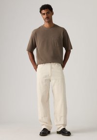 Καφέ κοντομάνικη μπλούζα με τσέπη, σε συνδυασμό με εκρού παντελόνι με λευκές ρίγες. Το μοντέλο φοράει μαύρα παπούτσια. Απλή, άνετη εφαρμογή.