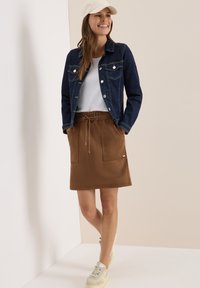 Veste en denim bleu foncé, t-shirt blanc et jupe à cordon de serrage marron avec deux poches avant, associée à des baskets beiges.