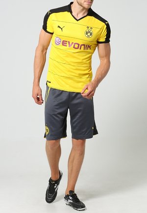 Mann trägt gelbes und schwarzes Borussia Dortmund Fußballtrikot mit grauer Shorts und schwarzen Fußballschuhen und geht nach vorne.