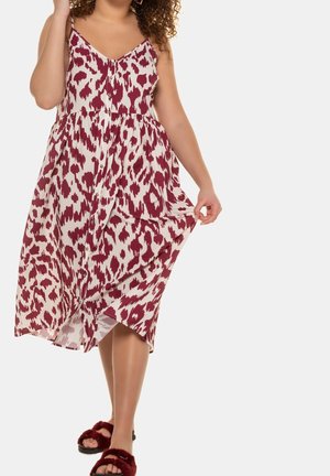 Frau trägt ein ärmelloses, weißes und burgunderfarbenes Midi-Kleid mit Animalprint und burgunderfarbene, pelzige Sandalen mit offenem Zehenbereich; hält mit einer Hand den Stoff des Kleides.