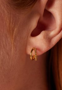 Gouden oorring met een gedraaide detail, met een gladde oppervlakte en cirkelvorm, gedragen op de oorlel tegen een huid achtergrond.